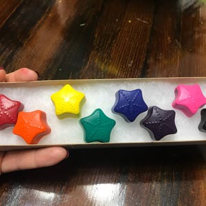 Könnte beinhalten: Eine Schachtel mit acht sternförmigen Buntstiften in einem Regenbogen aus Farben, darunter Rot, Orange, Gelb, Grün, Blau, Lila, Rosa und Schwarz.