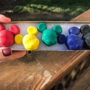 Op de afbeelding: Een set van vijf Mickey Mouse-vormige kleurpotloden in een kartonnen doos. De kleurpotloden zijn rood, geel, groen, blauw en zwart.
