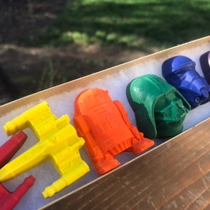 Puede incluir: Un conjunto de seis crayones con temática de Star Wars en una caja de cartón. Los crayones tienen forma de personajes y vehículos del universo de Star Wars, incluyendo Darth Vader, un Stormtrooper, R2-D2 y un caza X-Wing. Los crayones están dispuestos en un patrón de arcoíris, con rojo, naranja, amarillo, verde, azul y morado.