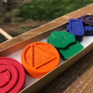 Puede incluir: Una caja de seis crayones de cera de colores en una variedad de formas y diseños. Los crayones están dispuestos en una fila, con el crayón rojo a la izquierda y el crayón morado a la derecha. Los crayones están todos en una caja con un forro blanco.