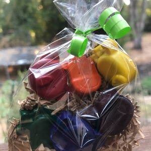 Puede incluir: Una bolsa transparente de celof&aacute;n atada con una cinta verde, llena de ceras de colores con forma de animales. Las ceras son rojas, naranjas, amarillas, verdes, azules y moradas. Las ceras est&aacute;n dentro de virutas de papel marr&oacute;n.