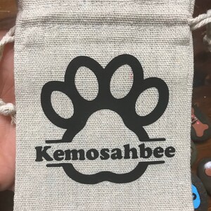 May include: A small, beige canvas bag with a black paw print and the name "Kemosahbee" printed on it. The bag has a drawstring closure and is perfect for holding small items.
