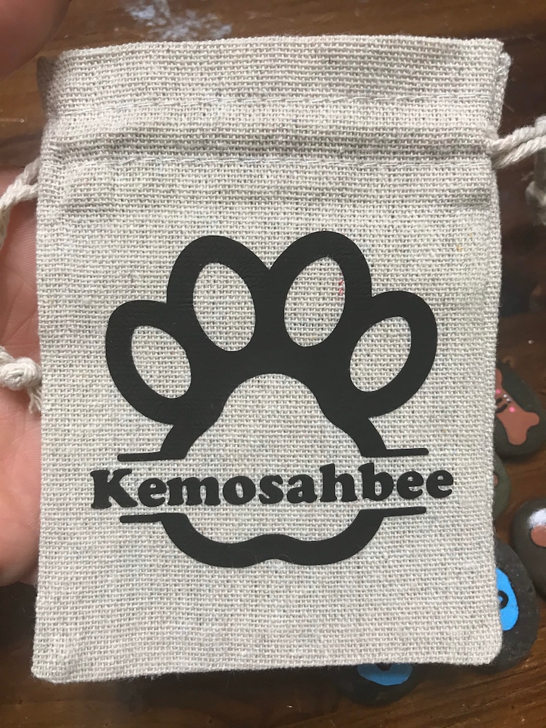 May include: A small, beige canvas bag with a black paw print and the name "Kemosahbee" printed on it. The bag has a drawstring closure and is perfect for holding small items.