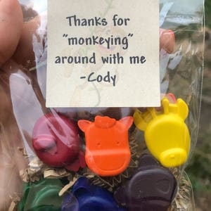 Puede incluir: Una bolsa transparente llena de crayones con forma de animales de varios colores. Los crayones son rojos, naranjas, amarillos, verdes, azules y morados. Una peque&ntilde;a tarjeta dice: "Gracias por 'monear' conmigo - Cody."