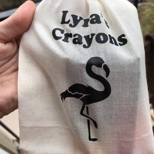 May include: A cream-coloured drawstring bag with the words "Lyra's Crayons" in black. A black flamingo silhouette is printed on the bag. The bag is held by a person.