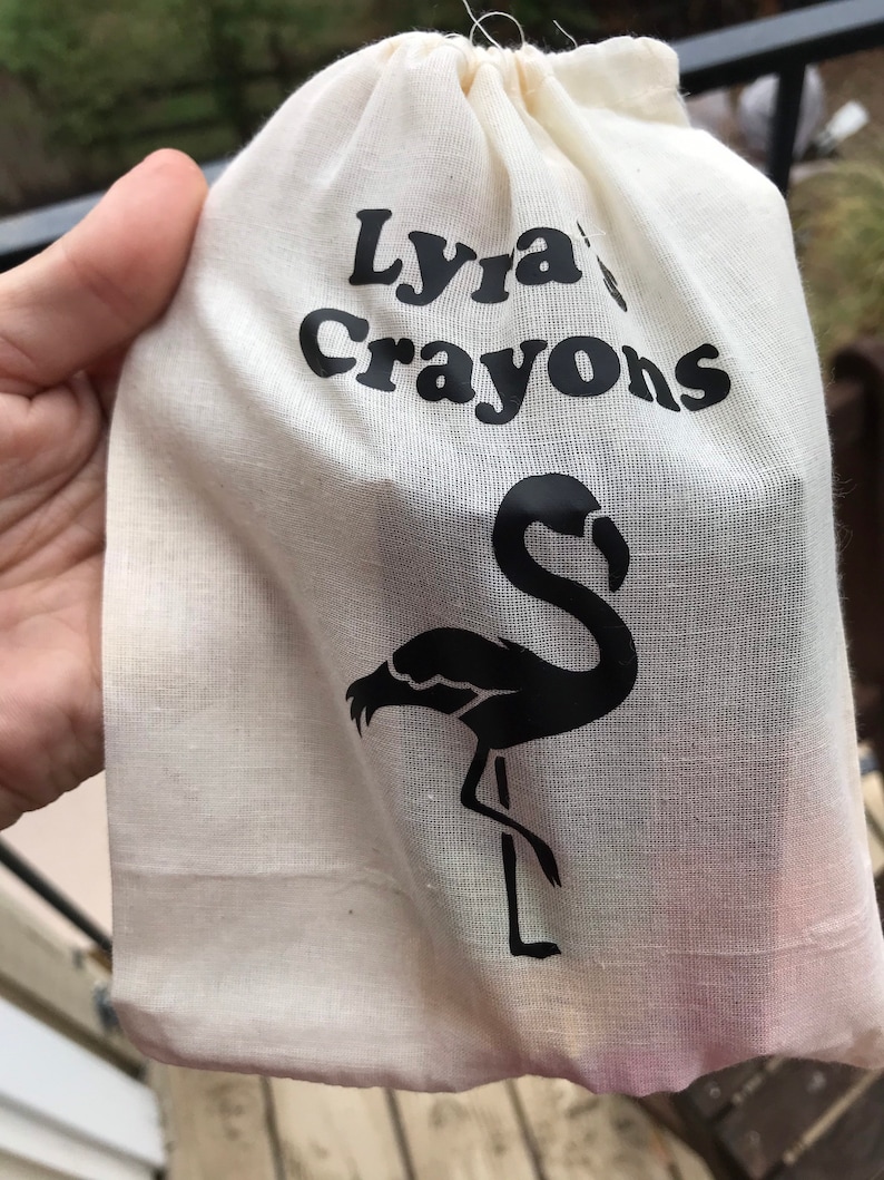 May include: A cream-coloured drawstring bag with the words "Lyra's Crayons" in black. A black flamingo silhouette is printed on the bag. The bag is held by a person.