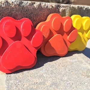 May include: Three paw print shaped molds, one red, one orange, and one yellow. The molds are made of a flexible material and are designed for making paw prints in concrete.