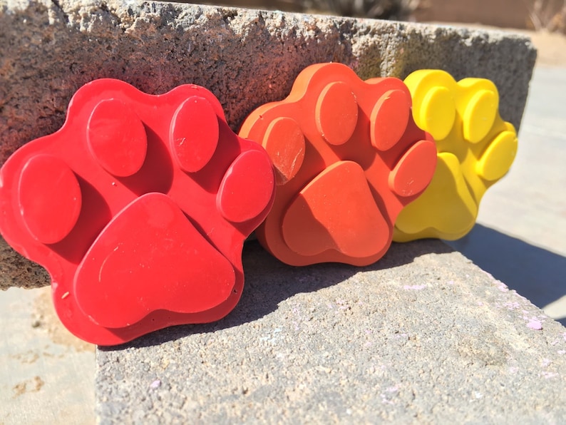 May include: Three paw print shaped molds, one red, one orange, and one yellow. The molds are made of a flexible material and are designed for making paw prints in concrete.