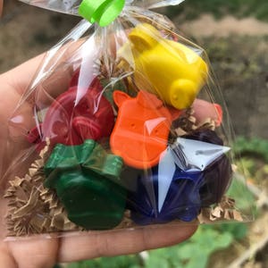 Puede incluir: Una bolsa transparente de celof&aacute;n atada con una cinta verde contiene ceras de colores con forma de animales. Las ceras son de varios colores, incluyendo rojo, naranja, amarillo, verde, azul y morado. La bolsa est&aacute; llena de papel marr&oacute;n triturado.