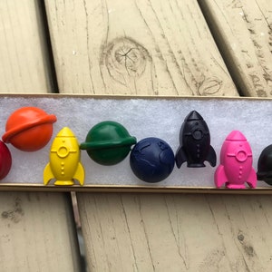 Peut inclure: Un ensemble de huit crayons en forme de planètes et de fusées. Les crayons sont dans une boîte doublée de feutre blanc. Les crayons sont rouge, orange, jaune, vert, bleu, rose et noir.