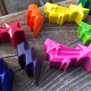May include: A set of eight colorful crayons shaped like shooting stars. The crayons are made of wax and have a smooth, glossy finish. The colors include red, orange, yellow, green, blue, purple, pink, and black.