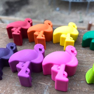 May include: A selection of vibrant flamingo-shaped bottle stoppers in shades of red, orange, yellow, green, purple, pink, and light green. The stoppers are made of a solid material and are displayed on a wooden surface.