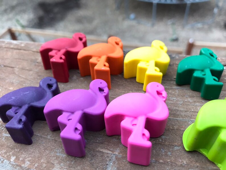 May include: A selection of vibrant flamingo-shaped bottle stoppers in shades of red, orange, yellow, green, purple, pink, and light green. The stoppers are made of a solid material and are displayed on a wooden surface.