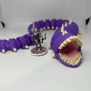 May include: A purple articulated worm figurine with cream-colored spikes and an open mouth revealing a pink interior, positioned next to a small, painted humanoid archer miniature standing on a circular base with a brick-patterned floor.