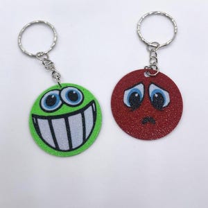 May include: Two keychains with round, glitter-covered faces. One face is green with a wide smile and large eyes. The other face is red with a frown and sad eyes.