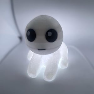 May include: A white, glowing figurine of a cartoon creature with large black eyes and a small mouth. The figure has a rounded head and a body with three stubby, glowing arms. The overall effect is soft and ethereal.