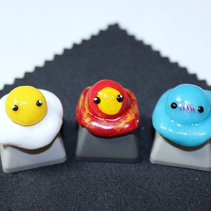 May include: Three keycaps with cute faces. The keycaps are shaped like a fried egg, a red mushroom, and a blue blob. The keycaps are on a black surface.