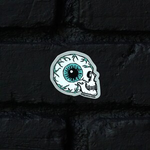 Pode incluir: Um adesivo de caveira branca com um olho azul e veias pretas. O adesivo está em uma parede de tijolo preto.