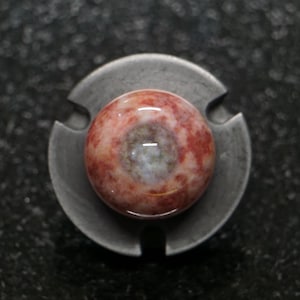 May include: A close-up of a decorative object featuring a round, polished stone with red and white marbling, set in a gray metal base. The stone has a central, lighter-colored area. The metal base has a three-pronged design.