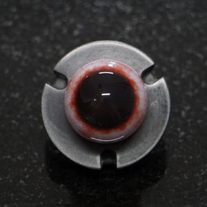 May include: A realistic glass eye with a dark brown iris and a red and white sclera. The eye is mounted on a silver metal base.