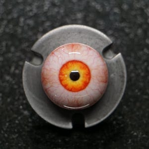 Puede incluir: Un ojo protésico detallado y realista con un iris amarillo y negro, rodeado por una esclerótica blanquecina con venas rojas. El ojo está montado en un soporte metálico gris, creando un contraste llamativo.
