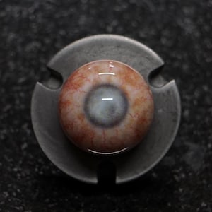 Puede incluir: Un ojo protésico realista con un iris detallado y vasos sanguíneos visibles, fijado en una base metálica de tres puntas. El ojo presenta un iris azul claro y está rodeado de venas rojas.