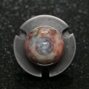 May include: A round, multi-colored stone cabochon set in a gray metal finding. The stone features swirls of red, brown, green, and blue hues. The metal setting has a circular shape with three evenly spaced cutouts.