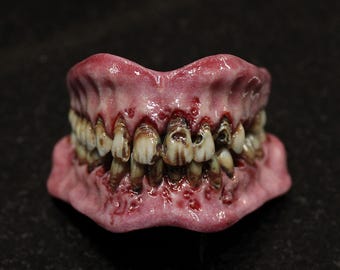 Puede incluir: Un modelo detallado e inquietante de una dentadura, con una línea de encías rosada y numerosos dientes que muestran signos de deterioro. Los dientes están manchados y dañados, con signos visibles de podredumbre y descomposición. Un accesorio para Halloween o temas de terror.