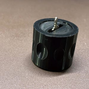 May include: A black cylindrical object with three circular cutouts and a silver metal piece inserted in the top. The object is sitting on a brown surface. The cylindrical object appears to be a pencil sharpener.