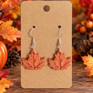 May include: A pair of dangle earrings featuring orange maple leaf charms with a glittery texture. The leaves are attached to silver-toned metal fishhook wires and are displayed on a rectangular brown cardboard jewelry display card.
