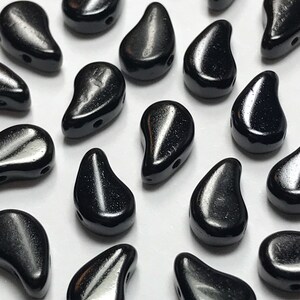 May include: Close-up of numerous black teardrop-shaped beads. Each bead has a glossy finish and a small hole for stringing. The beads are scattered on a white surface, creating a pattern of light and shadow.