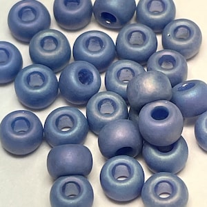 May include: A collection of blue-purple round beads with a matte finish. Each bead has a large, central hole. The beads are scattered on a white surface, creating a visually appealing arrangement. These beads are suitable for jewelry making and crafting projects.