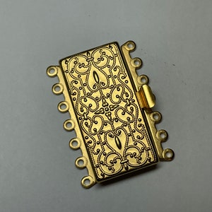 May include: Gold-toned metal clasp with an ornate design and multiple loops for attaching jewelry or other items.