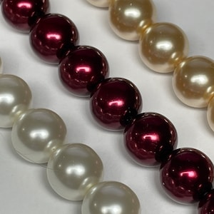 May include: Close-up of three strands of large, round beads. One strand features creamy white beads, another has deep burgundy beads, and the third strand has light beige beads. The beads are strung together, creating a classic jewelry design.