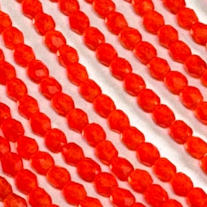May include: A close-up of multiple strands of bright orange faceted glass beads. The beads are arranged in rows on a white surface.