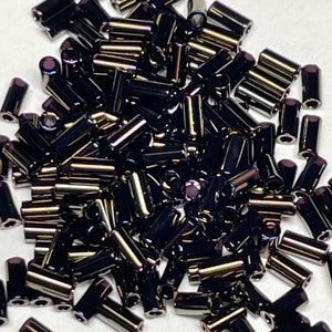 May include: A pile of black iridescent glass beads, shaped like small cylinders. The beads have a metallic sheen and are scattered on a white surface.