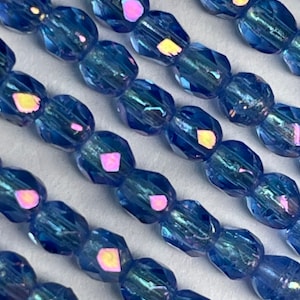 May include: Close-up of rows of faceted, iridescent blue beads. The beads have a deep blue color with flashes of pink and yellow. The beads are strung together, suggesting they are for jewelry making or crafts.