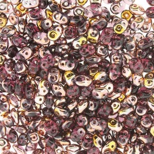May include: A close-up of a pile of small, iridescent glass beads. The beads are a mix of purple and gold, and they have a slightly irregular shape.