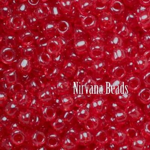 May include: Close-up of numerous small, transparent red glass beads. The beads are scattered, creating a textured surface. The text "Nirvana Beads" is printed in white across the image.