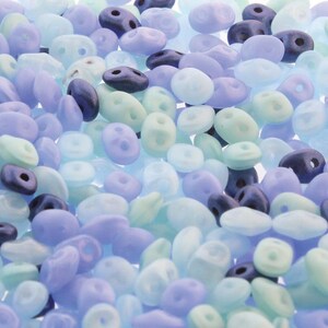 May include: A collection of small, oval-shaped two-hole beads in a mix of matte and translucent finishes. The color palette consists of various shades of light blue, periwinkle, soft lavender, mint green, and dark navy blue scattered together.
