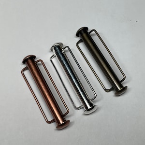 May include: Three metal slider buckles in different finishes: copper, silver, and antique brass. Each buckle has a rectangular shape with a cylindrical center and two small loops on either side.