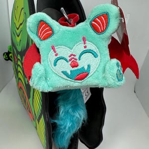 Puede incluir: Un murciélago de peluche de color turquesa con alas rojas y una cara sonriente. El murciélago está unido a una cabeza de monstruo de peluche negra y verde.