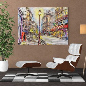 Pode incluir: Uma pintura que representa uma cena de rua parisiense com um poste de luz no centro. Os edif&iacute;cios s&atilde;o pintados em um estilo colorido e abstrato. A pintura est&aacute; pendurada em uma parede acima de uma poltrona e um pufe brancos.