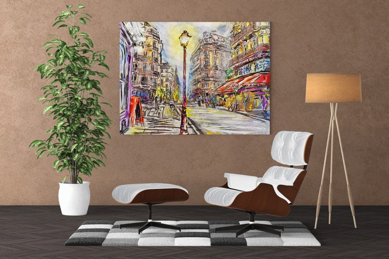 Pode incluir: Uma pintura que representa uma cena de rua parisiense com um poste de luz no centro. Os edif&iacute;cios s&atilde;o pintados em um estilo colorido e abstrato. A pintura est&aacute; pendurada em uma parede acima de uma poltrona e um pufe brancos.