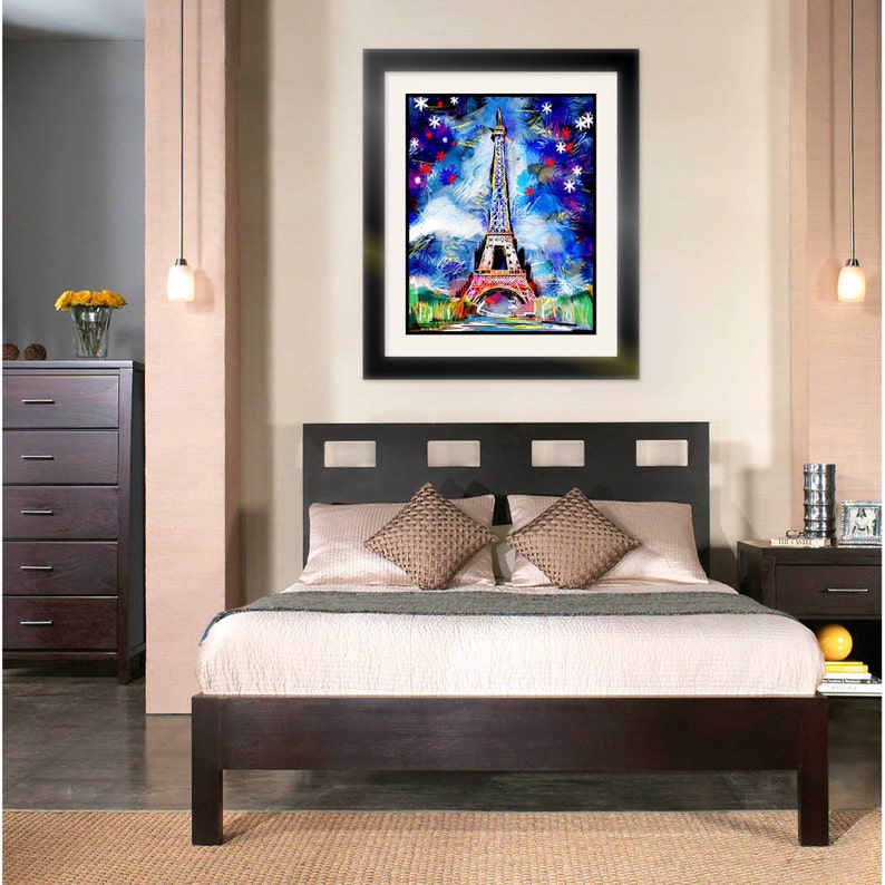 May include: A framed painting of the Eiffel Tower in Paris, France. The painting is done in a colorful abstract style with a blue background and white snowflakes. The painting is hanging on a wall in a bedroom with a dark wood bed frame and white bedding.