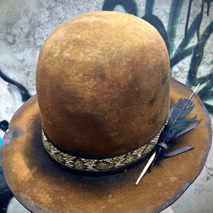 May include: A brown fedora hat with a wide brim and a rounded crown. The hat features a patterned band and is adorned with a black feather and a small, light-colored stick. The hat has a distressed, vintage look.