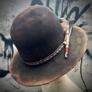 May include: A dark brown fedora hat with a distressed finish. The hat features a decorative braided band and is adorned with feathers. The brim has a slightly worn appearance, adding to its vintage aesthetic.