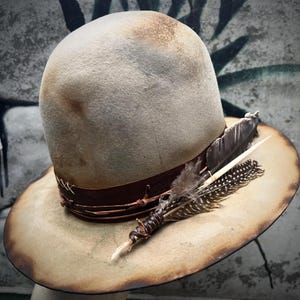 May include: A distressed-look fedora hat with a light brown crown and brim. The hat features a dark brown leather band with barbed wire detailing and is adorned with decorative feathers. The brim has a darker, aged appearance.