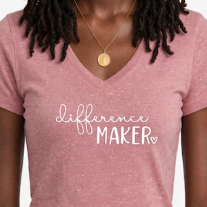 May include: A heather rose V-neck t-shirt featuring the white text "difference MAKER" with a small heart icon. The shirt has short sleeves and a textured fabric appearance. A gold-toned pendant necklace rests against the neckline of the shirt.