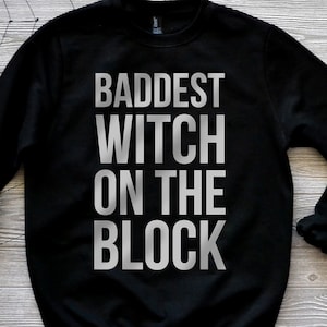 May include: A black long-sleeve crewneck sweatshirt featuring the text "BADDEST WITCH ON THE BLOCK" printed in large, bold, light gray capital letters across the center of the chest. The garment has a ribbed collar, cuffs, and hem.